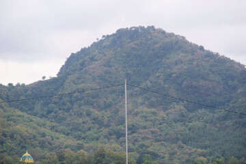 A green forested mountain peak under a cloudy sky, showcasing natural tropical scenery.