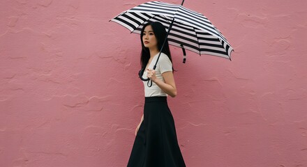 Fototapeta premium Woman walks gracefully holding a black and white striped umbrella against a solid pink textured wall during a light afternoon shower.