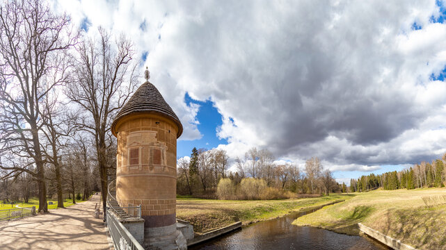 Panoramic view of Pavlovsk Park: Slavyanka River meanders past green meadows, forests, and the iconic thatch-roofed Peel Tower pavilion. Sunny May serenity. Pavlovsk, St. Petersburg, Russia. 