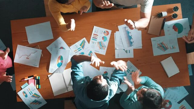Top view of smart people throw paper and giving high five at meeting table with financial document and stock market analysis paper scattering around. Aerial view of team celebrate project. Symposium.