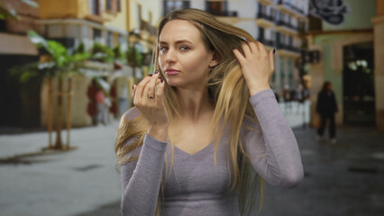 Fototapeta premium Woman examining long hair outdoors on a sunny urban street surrounded by buildings and palm trees with a thoughtful expression.