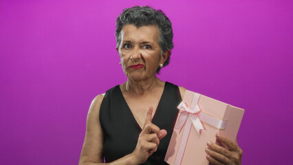 Senior woman with grey hair examines a pink gift box against a vibrant pink background, expressing...