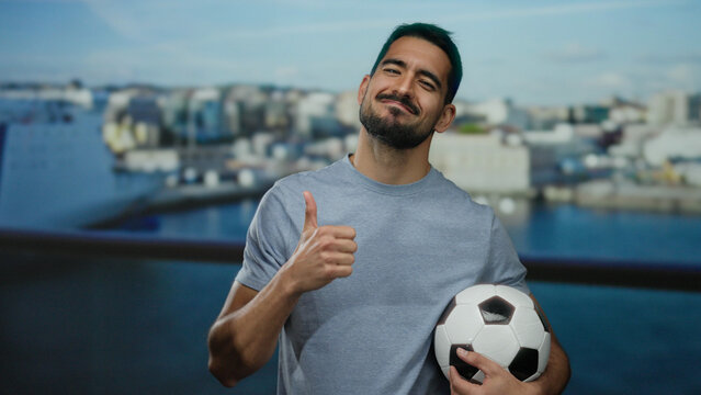 Young man smiling confidently by seaside holding soccer ball with thumbs up in casual shirt outdoors showcasing happiness and leisure with vibrant backdrop of port.