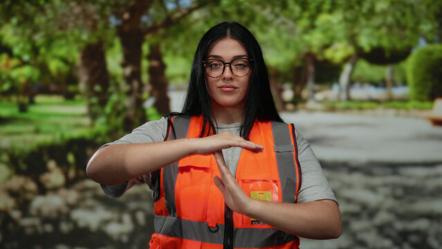 Woman volunteer wearing glasses and an orange vest makes a timeout gesture in a sunny park setting, featuring lush green trees and a serene outdoor landscape.