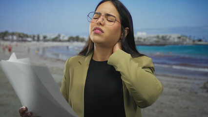 Woman rubbing neck looks stressed on a beach with a young latin hispanic presence by a seaside outdoors holding a paper.