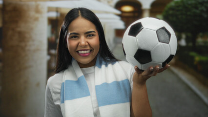 Woman smiling happily outdoors holding soccer ball in sunny courtyard wearing blue scarf enjoying...