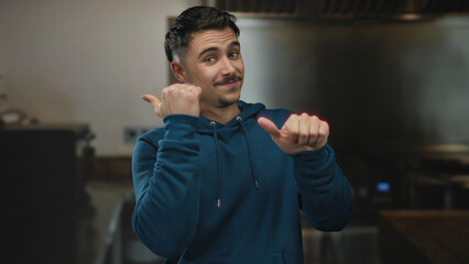 Young hispanic man in blue hoodie smiling and pointing thumbs sideways in kitchen setting offering...