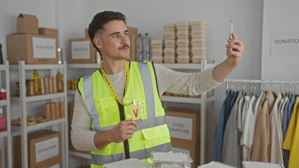 Young hispanic man in reflective vest takes a selfie at an indoor charity center filled with donations, surrounded by organized boxes and clothing racks.