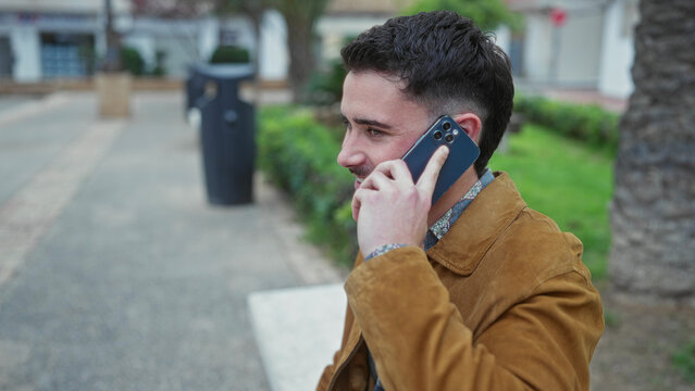 Young man talking on smartphone wearing a brown jacket outdoors in a sunny park setting, showcasing modern urban lifestyle and communication.