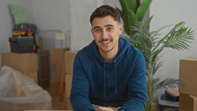 Young man in new home living room, indoors, wearing hoodie, smiling, talking to camera, surrounded by moving boxes, plants, and unpacked items, hispanic ethnicity.