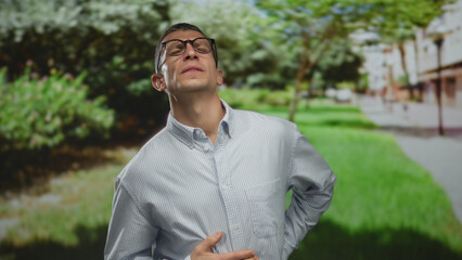 Caucasian man in glasses experiencing stomach pain while standing in a sunny green park with a...