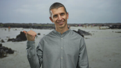 Man smiles on a serene beach pointing sideways against a backdrop of sea and rocks, wearing a grey shirt and enjoying a tranquil seaside setting