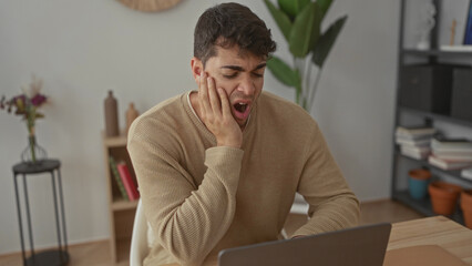 Man holds hand to face yawning at laptop while young hispanic student works at home male computer desk.