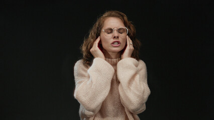 Woman covers her ears to block noise on isolated black background wearing glasses and sweater...