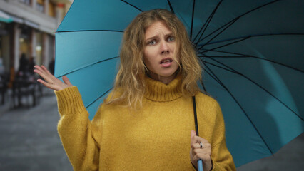 Woman holding umbrella displays a confused expression on street, hair dyed blonde beside a blue...