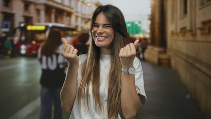Young hispanic woman smiling making money gesture on busy city street during daytime with blurred...