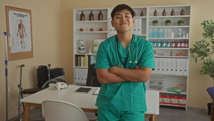 Obraz premium Young chinese doctor in green scrubs with arms crossed in a clinical room adorned with medical supplies, conveying a professional yet relaxed atmosphere.