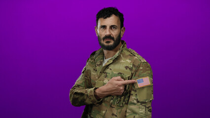 Hispanic man with beard in military uniform points to american flag patch against a vibrant purple...