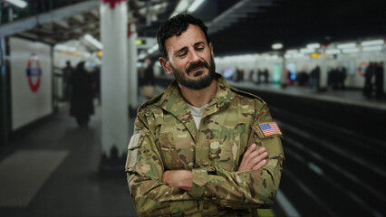 Fototapeta premium Man in military uniform standing in an underground station with arms crossed, displaying a thoughtful expression amid blurred commuters and subway signage, highlighting urban transit mood.