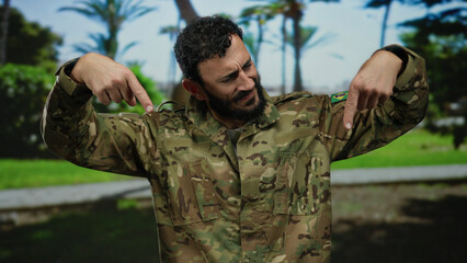 Obraz premium Hispanic man with beard wearing military uniform points downward in a sunny green park with trees in the background.