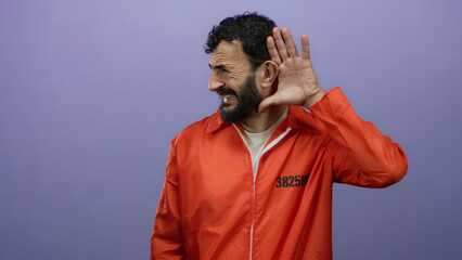 Hispanic man with beard wearing prison jumpsuit gestures listening against purple background.