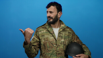 Hispanic man with beard in military uniform holding helmet against blue background, exhibiting a...
