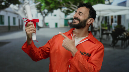 Hispanic man in orange prison jumpsuit smiles outdoors on a street while holding and pointing at a degree scroll with a red ribbon, symbolizing success and achievement.