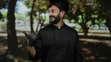 Man chef with beard smiling outdoors in a green park, wearing black uniform and gloves, surrounded...