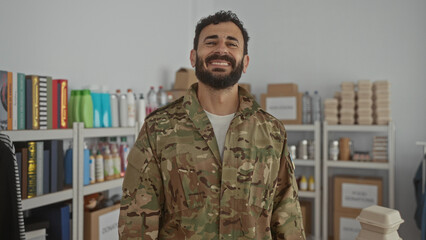 Hispanic man with beard in military uniform stands indoors in a volunteer room with supplies and...