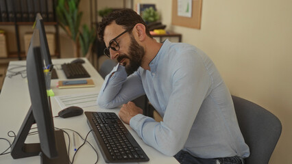 Hispanic man with beard, tired and resting head on hand at office desk with computer, reflecting...