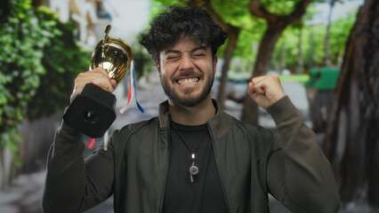 Young man celebrating victory with trophy outdoors on a vibrant street showing overwhelming joy and...