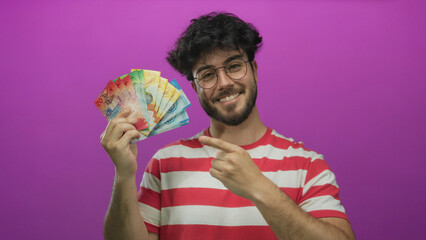 Young hispanic man with beard in striped shirt smiling and pointing at swiss francs against vivid...