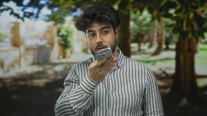Young hispanic man with beard using smartphone outdoors on a street, wearing striped shirt and appearing focused amidst a natural background of trees and sunlight.