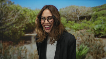 Woman laughing outdoors in park with lush greenery wearing glasses and blazer conveying joyful and...