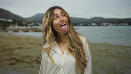 Young blonde woman with long wavy hair sticks tongue out on a sandy beach shore; silliness fun joy...