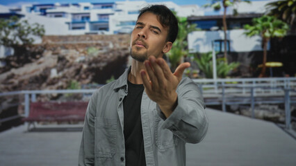 Young man in city street making come here gesture with serious expression outdoors under bright sky.