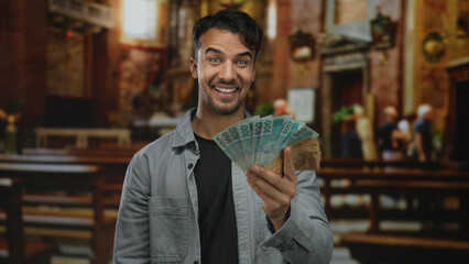 Fototapeta premium Young man in church holding brazilian money smiling indoors showcasing wealth and happiness in a religious setting with vibrant surroundings.