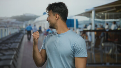 Fototapeta premium Young hispanic man pointing thumb back on cruise boat outdoor wearing casual clothes under cloudy sky highlighting relaxed atmosphere.