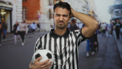 Fototapeta premium Young hispanic referee in a striped shirt holding a soccer ball on a busy city street appears frustrated amid an urban backdrop.