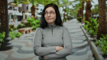 Woman with brunette hair smiling outdoors wearing glasses and a gray sweater on a palm-lined street with green plants creating a lively urban backdrop.