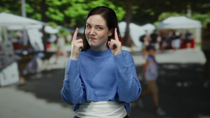 Woman outdoors in blue sweater points upward on a sunny day with market stalls in the background,...