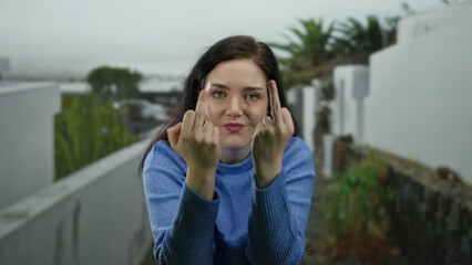 Woman in blue sweater makes expressive gesture outdoors on urban street with greenery in...