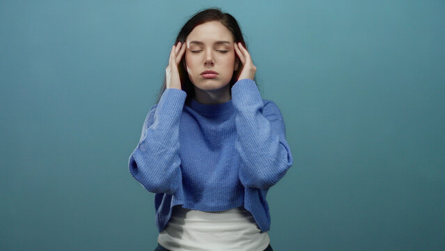 Woman with brunette hair feeling stressed in front of a blue background struggles with headache wearing a blue sweater capturing emotion and tension vividly.
