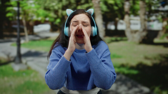 Woman wearing headphones and gesturing emphatically in a green park setting, conveying an emotional expression outdoors.