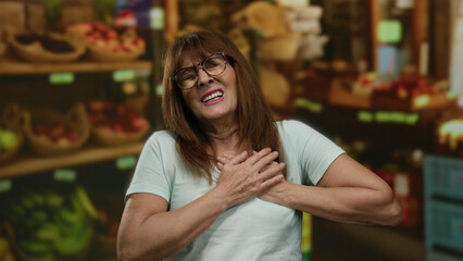 Fototapeta premium Hispanic senior woman clutching chest in fruit shop, expressing distress, surrounded by colorful produce outdoors, evoking concern and urgency in a lively market setting.