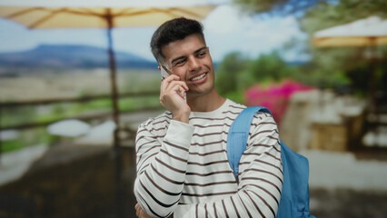 Young man on phone call standing on a city terrace, wearing striped shirt and backpack, with...