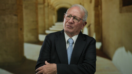 Senior man in a suit stands in an ancient university hallway reflecting on past achievements with a...