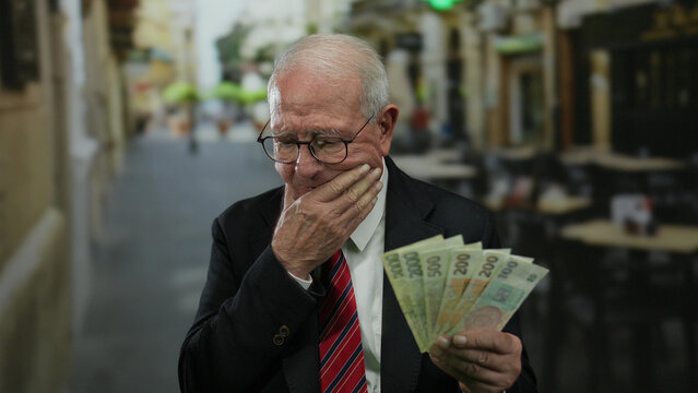 Senior man in a business suit ponders while holding czech koruna banknotes on an outdoor restaurant terrace on the street.
