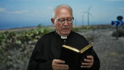 Senior priest man holding a book in an outdoor field with windmills in the background, illustrating...