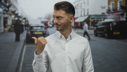 Man in white shirt standing on a narrow cobblestone city street points thumb to blurred cars and...